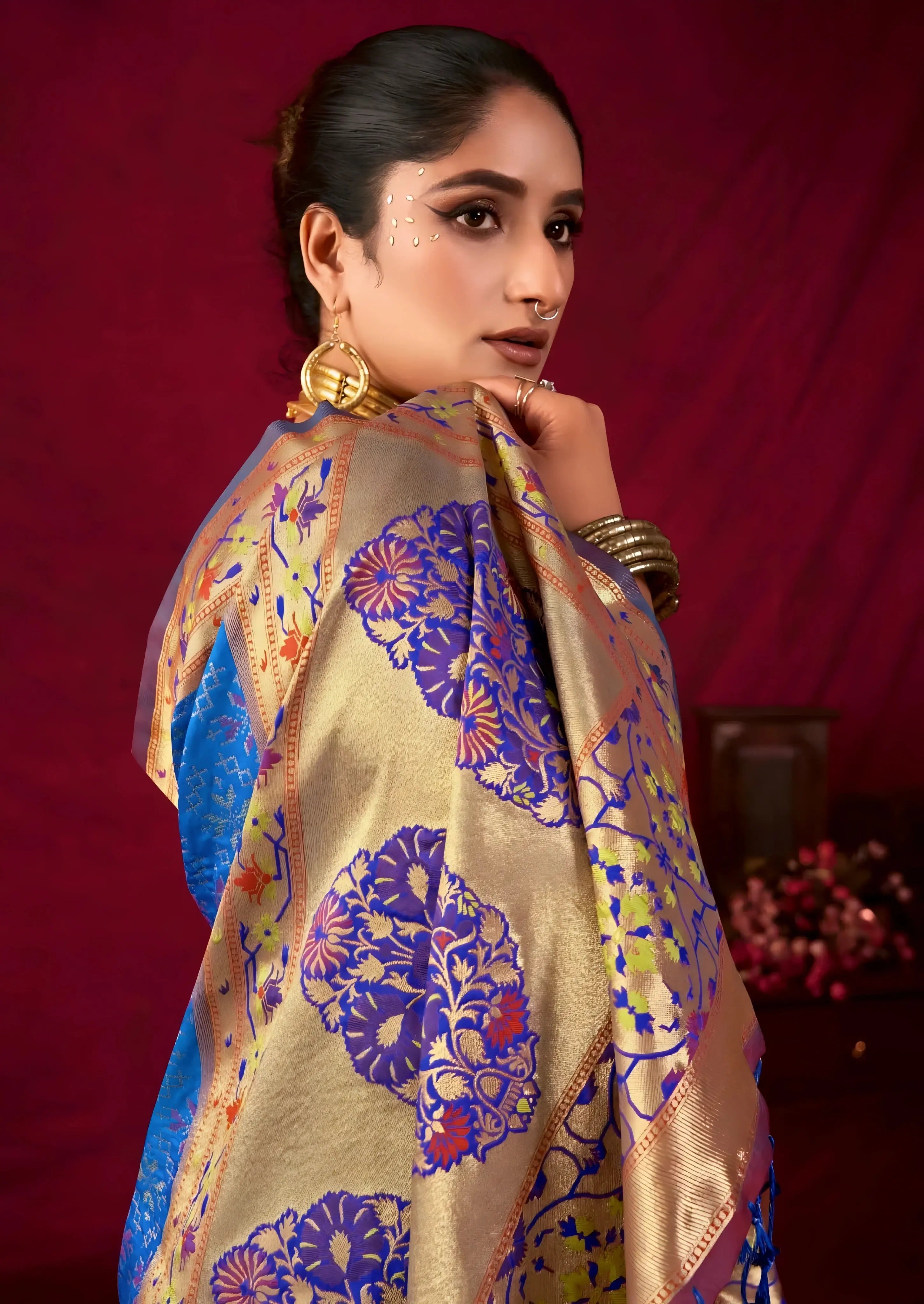 A woman models a luxurious Royal Blue Soft Banarasi Silk Saree. The wide border and pallu feature dense gold Zari weaving with prominent purple accents and large traditional motifs. She wears a unique dual-toned (purple/mauve) blouse and a heavy gold Kundan choker necklace, posing indoors.